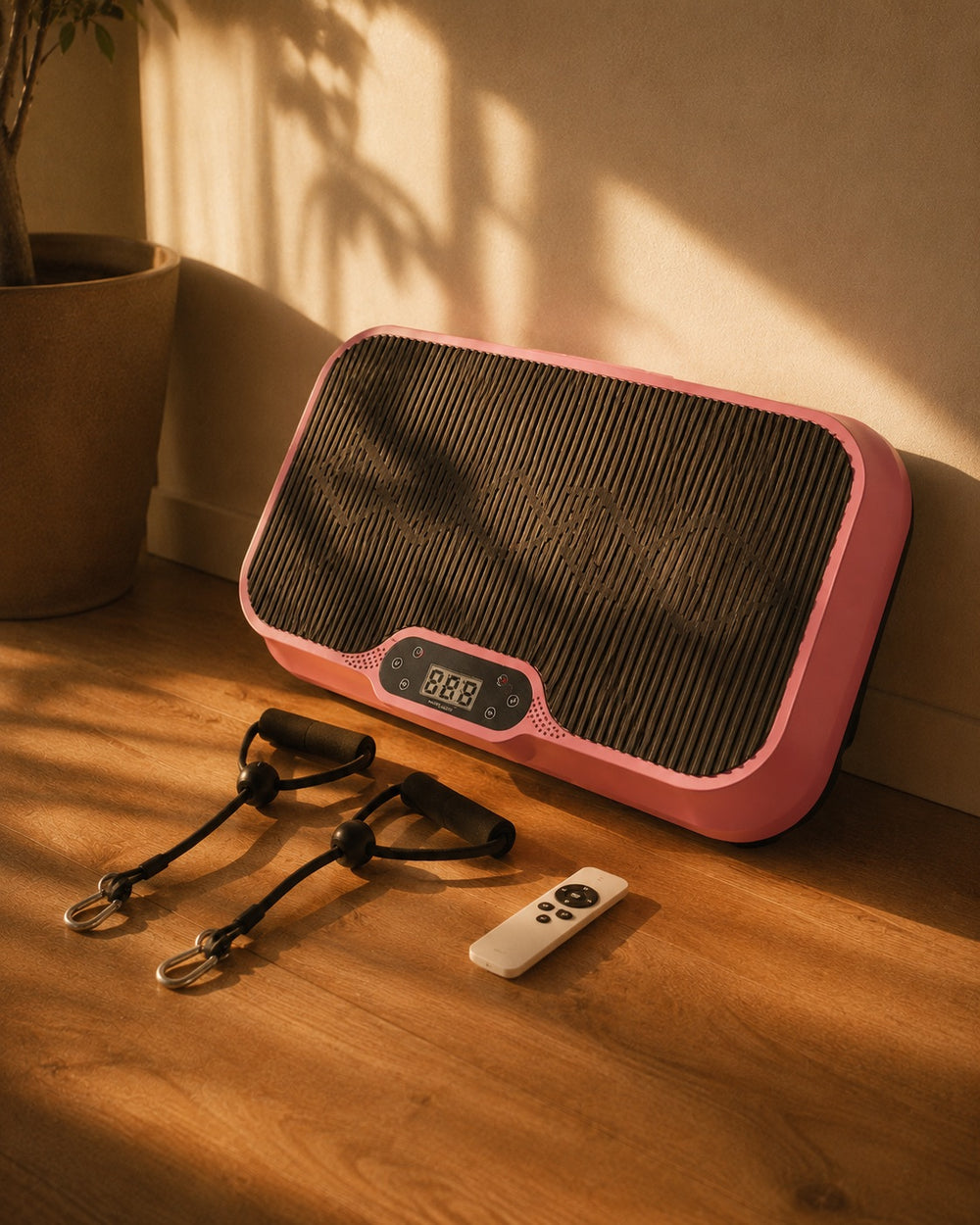 The pink Nuvé plate leaning against a wall with the remote and two resistance bands on the wood floor
