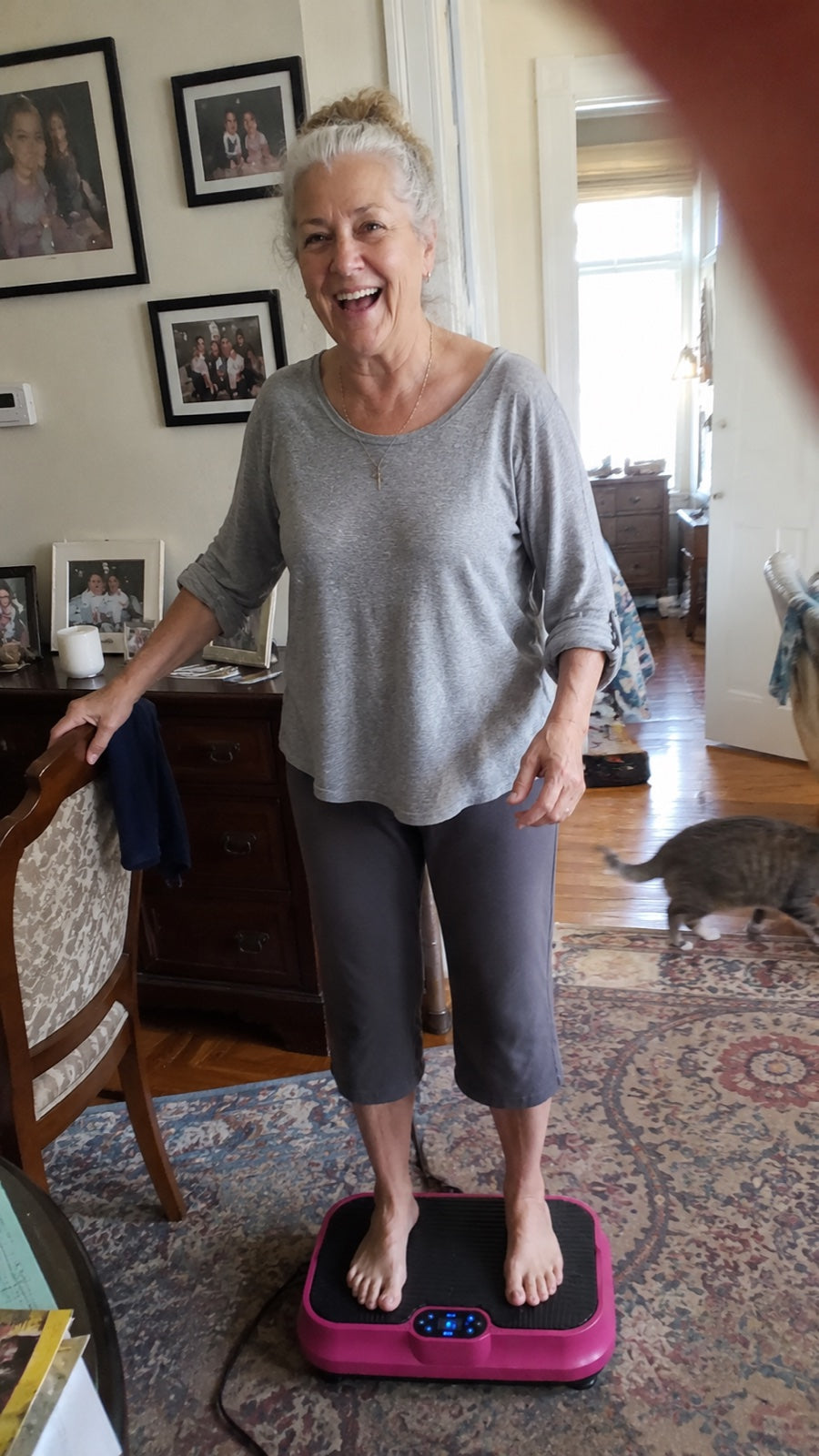 Woman standing barefoot on the pink Nuvé plate in her living room, laughing, her cat walking past